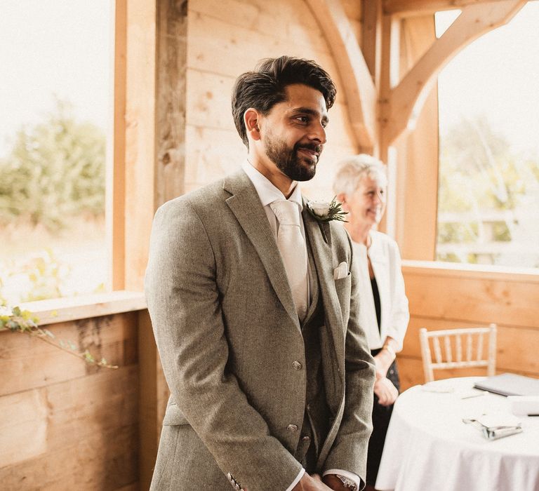 Groom in grey three piece suit, white tie and white floral buttonhole smiles as he waits for bride at Inkersall Grange Farm wedding ceremony