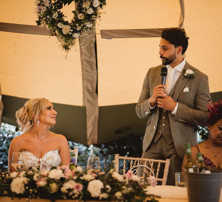 Bride in off the shoulder Enzoani wedding dress and curly wedding updo smiles at groom in grey three piece suit with white tie and buttonhole as they sit at top table during tipi wedding breakfast at Inkersall Grange Farm wedding