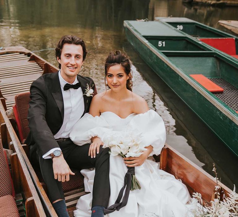 Stylish bride and groom in a tuxedo and Nortier Shallow wedding dress sitting on a boat at their Oxford wedding 