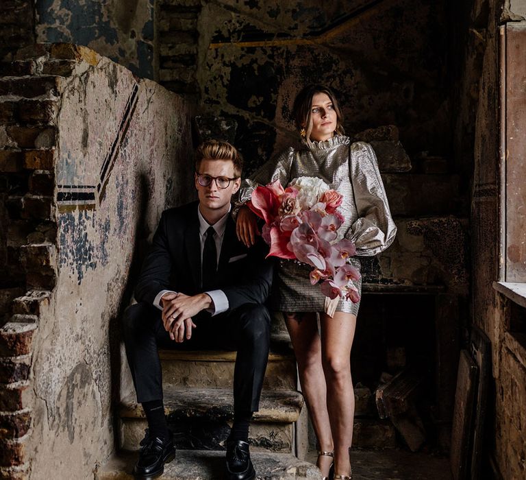 Cool Bride in gold platform heels with a bouquet of pink tonal flowers alongside Groom sitting on stone brick staircase in a dark suit industrial style wedding 