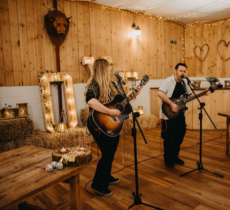 Two guitarists sing at microphones during rustic wedding ceremony in Lancashire with fairy lights, wooden tables and hay bales 