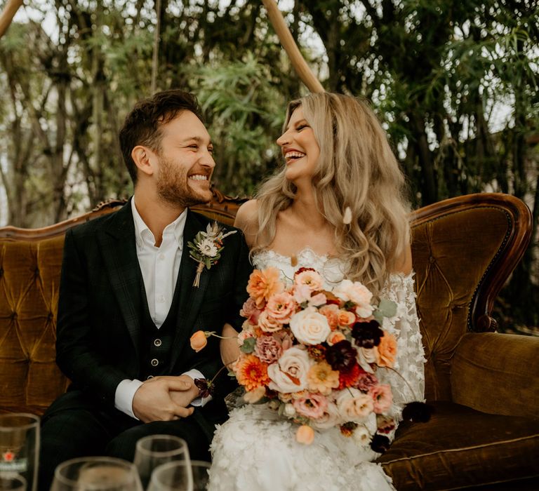 Bride in off the shoulder boho wedding dress holding white, pink and orange autumnal bridal bouquet sits on brown vintage sofa laughing with groom in dark suit and floral buttonhole