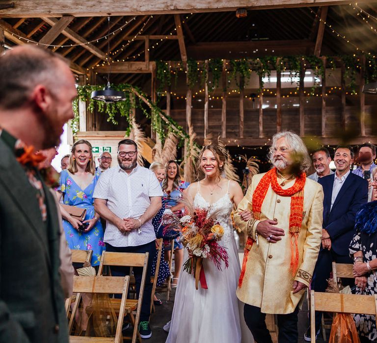 Bride in lace and tulle wedding dress holding mixed dried floral bouquet walks down the aisle arm in arm with wedding guest in yellow coat and orange scarf at festival inspired wedding