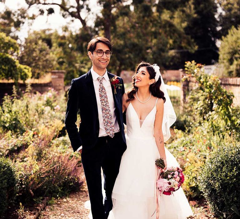 Bride in wedding dress with plunging neckline in feathered headband holding pink and red bridal bouquet walks through gardens with groom in dark suit and Gucci floral tie