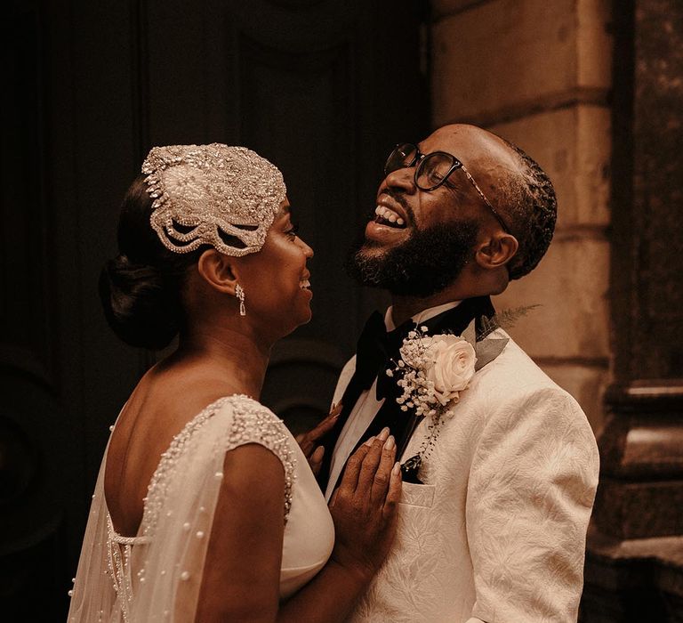 Groom in a white brocade tuxedo jacket laughing with his bride in a Pronovias wedding dress with pearl embroidered Watteau train 