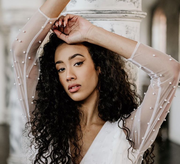 Black bride with naturally curly hair and winged eyeliner in a pearl embellished wedding dress with long sleeves by Story Of My Dress