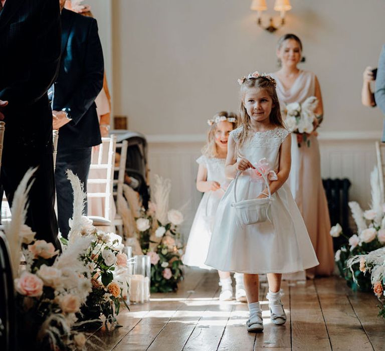 Flower girls in white dresses with flower crowns and baskets walking down the aisle