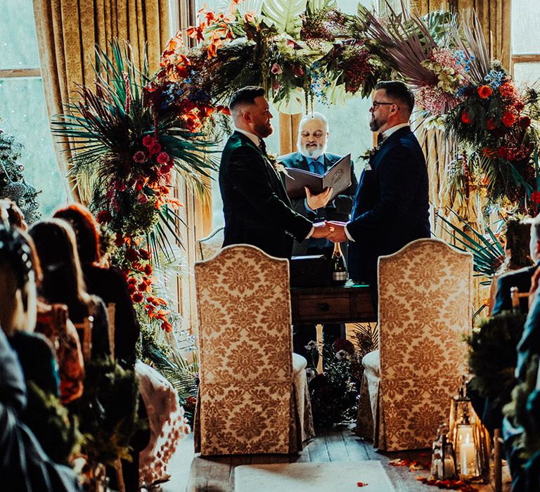 Grooms stand at the altar together in velvet suit jackets in front of damask patterned chairs and floral moongate