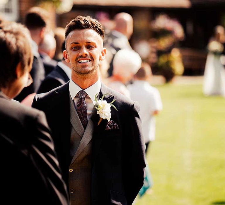 Groom in three piece suit with black suit jacket, grey waistcoat, paisley tie and white flower boutonnière waits to take his first look