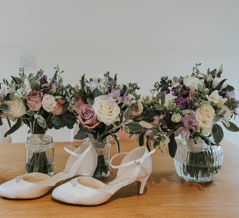 Bridesmaids bouquets resting in clear glass vases with purple flowers and sparkly low heeled white bridal shoes 