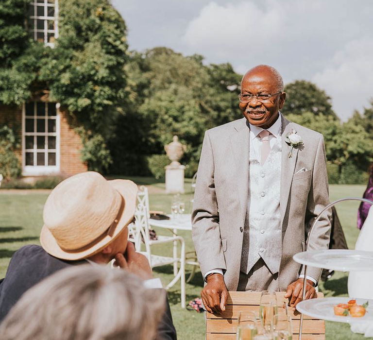 Father of the bride in grey suit and patterned waistcoat stands at outdoor tables for outdoor wedding