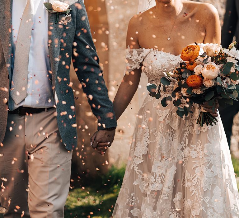 Groom in grey suit and pink tie holds hands with bride in off the shoulder floral lace wedding dress holding orang and white bouquet 