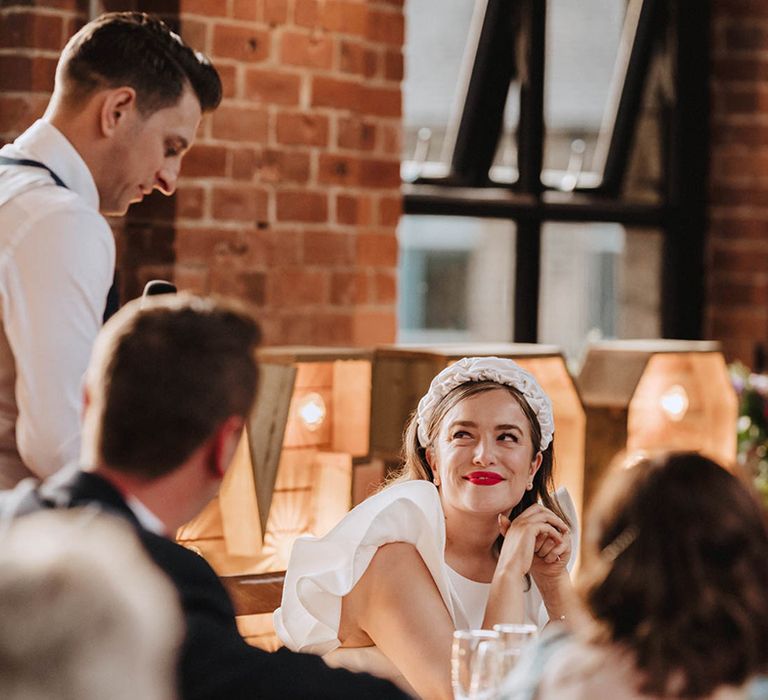 Groom in waistcoat reads out his speech as the bride smiles at him adoringly wearing red lipstick and large white headband 