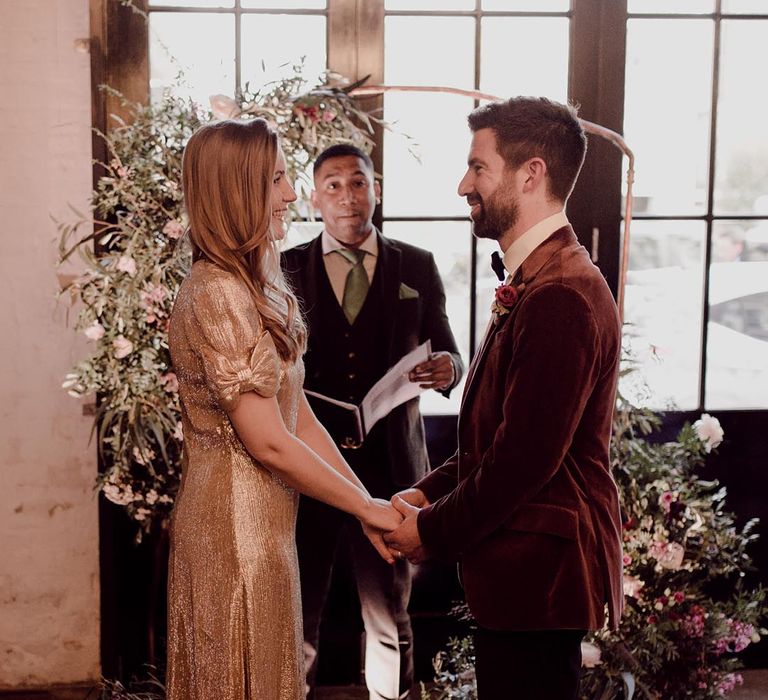 Groom in burgundy velvet suit jacket holds hands with bride in sparkly gold wedding dress with bow sleeves at the altar in front of frame with flower decor 