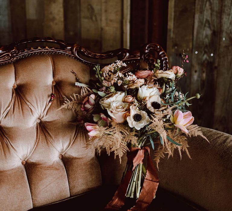 romantic pink rose, anemone and astilbe wedding bouquet tied with ribbon resting on an ornate sofa