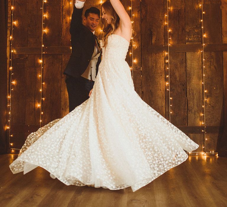 Groom in blue suit spins the bride around as they have their first dance in front of fairy light backdrop