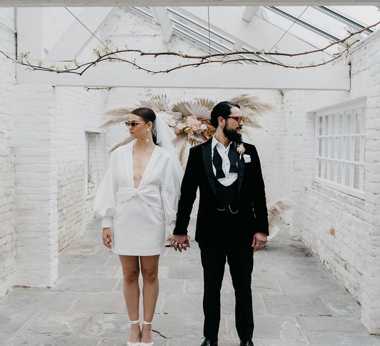 Stylish bride and groom in a tuxedo and short wedding dress with platform shoes, bow hair accessory and wedding sunglasses at Garthmyl Hall 