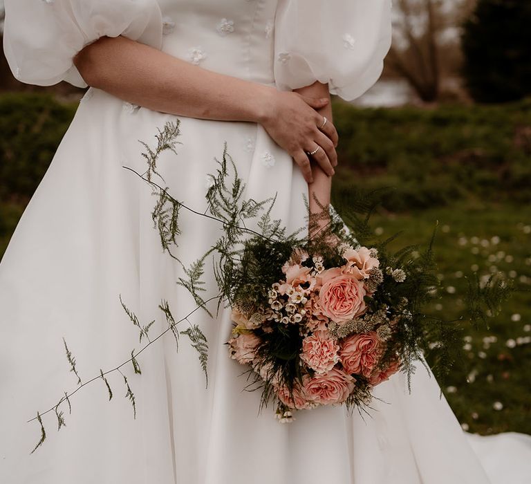 Bride in a strapless wedding dress with V front and puff sleeves holding a pink rose and carnation wedding bouquet 