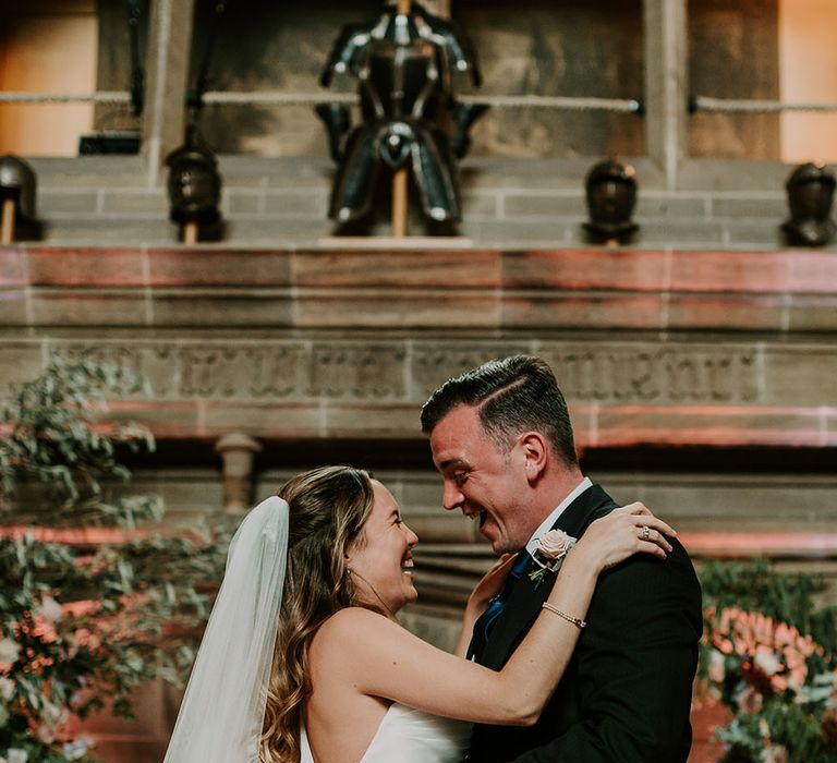 Bride wears princess wedding dress and floor-length veil whilst looking lovingly at her groom who wears morning suit and floral buttonhole