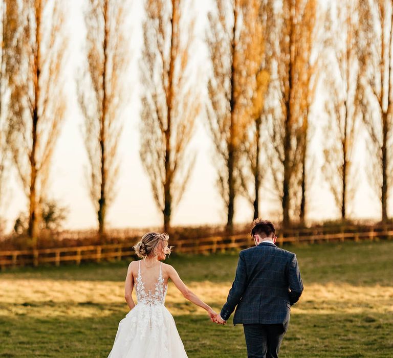 Bride in Madison James illusion back button wedding dress walking hand in hand with the groom in a blue suit 