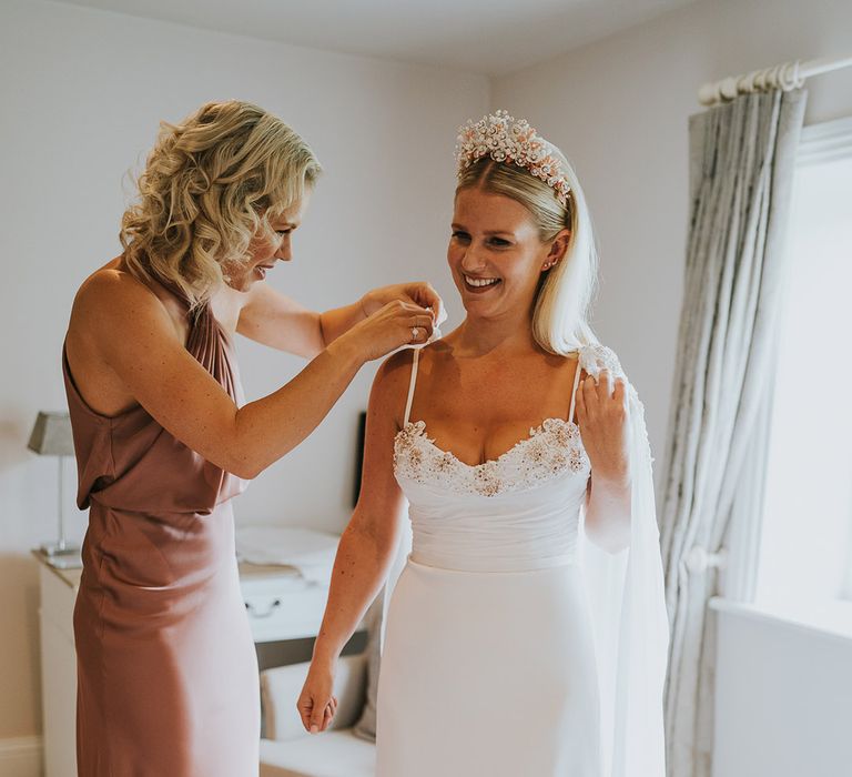 Bride in flower embellished wedding dress with large crown gets help from bridesmaid in dusty pink satin dress 