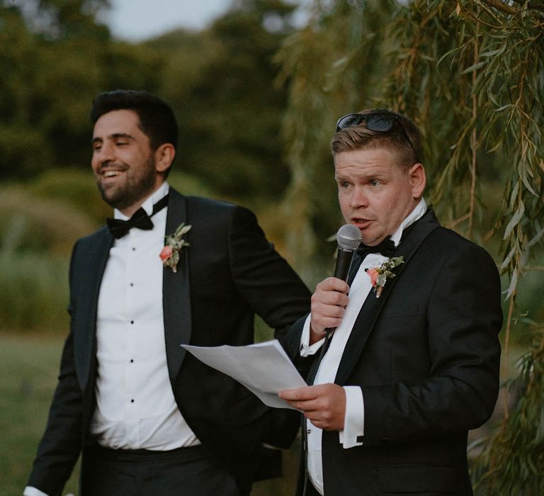 Groomsmen wear black tie and floral buttonholes whilst reading speech in outdoor wedding reception