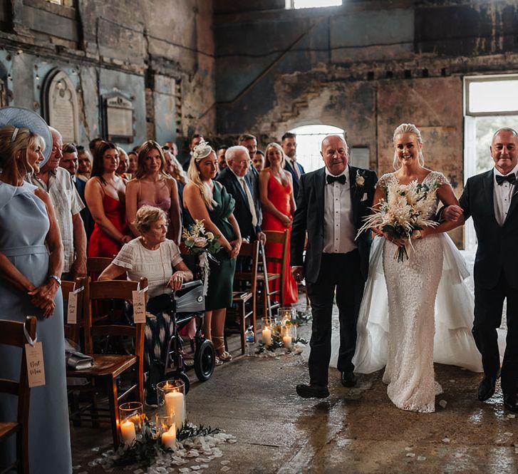 Bride holds pampas grass bouquet as she walks down aisle lined with pillar candles at The Asylum 