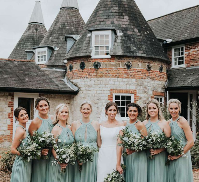 Bride in off the shoulder wedding dress and bridal party wearing teal bridesmaid dresses standing outside Bury Court Barn