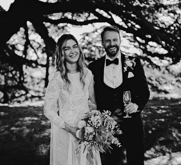 Groom in black tie drinking champagne with the bride in a long sleeve lace wedding dress holding a bouquet 