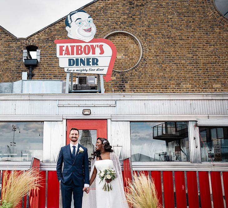 Bride wearing tulle cape with glitter embellishment & groom wearing three-piece suit stand in front of colourful diner on their wedding day 