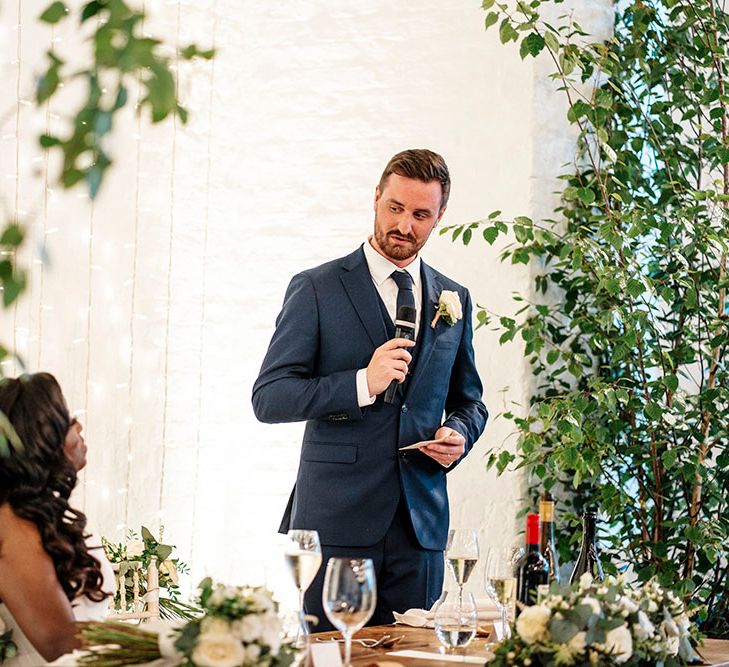 Groom gives speech during reception surrounded by green foliage 