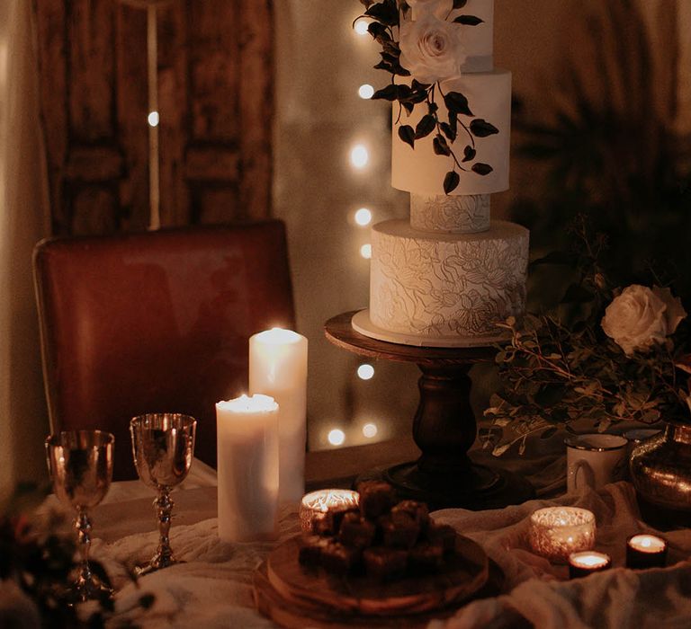 White iced textured wedding cake on a wooden cake stand with tea lights, pillar candles and golden goblets