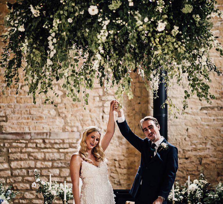 Foliage and white flower cloud hanging in the barn venue as the bride and groom raise their arms in celebration of their marriage 