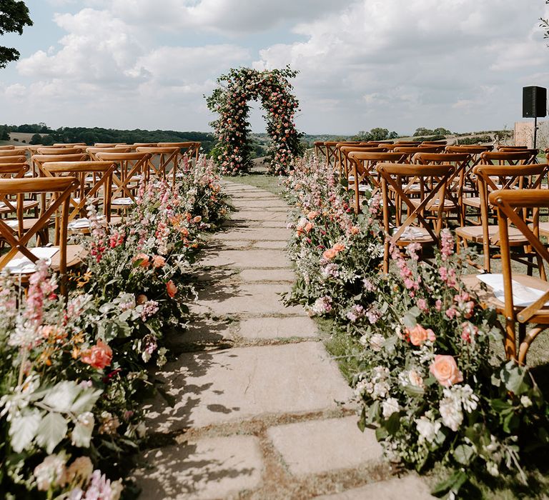 Wooden chairs line outdoor aisle complete with colourful floral arrangements and archway to the end of aisle