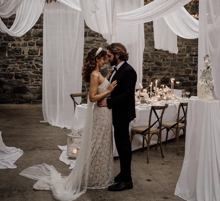 Bride in sleeveless lace pearl wedding dress and pearl headband with groom in black tux kissing amidst ceiling drapes decoration