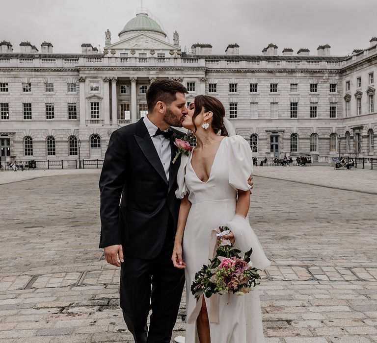 Bride in front slit wedding dress with puff sleeves embracing groom in dark tux sharing a kiss at Somerset House