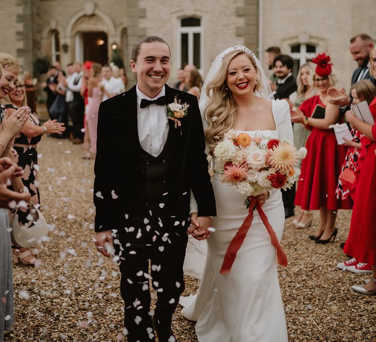 Bride in square neck long sleeve satin wedding dress with two tier veil and mixed floral bouquet with red ribbon holding hands with groom in black suit with floral boutonniere doing confetti walk