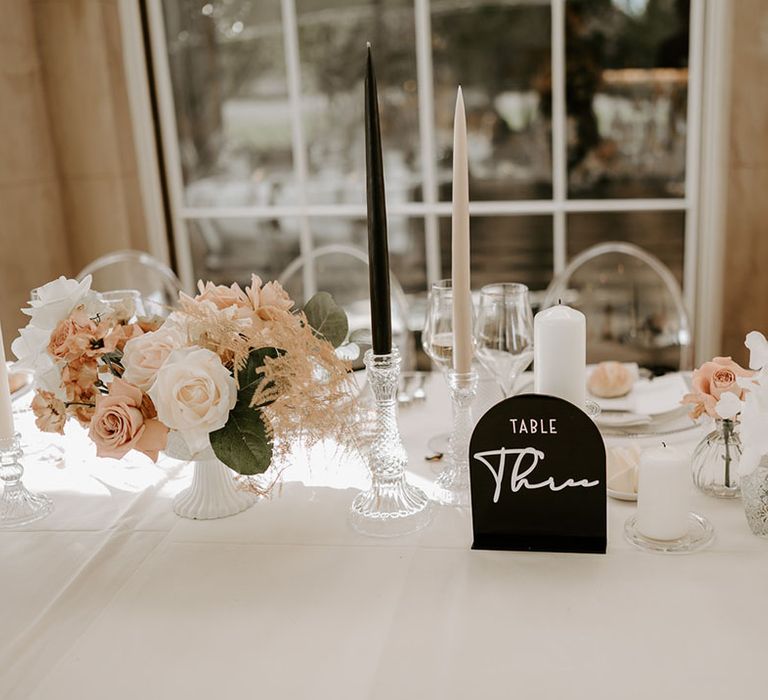 Pale pink and white wedding flower arrangements with a black table name sign and taper candles 