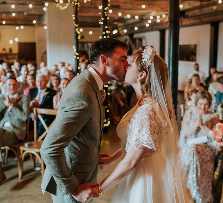 Groom in a grey tweed suit sharing his first kiss with the bride as a married couple with white flower headband and pearls 