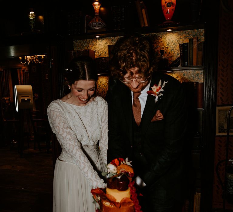 Bride in long sleeve lace vintage wedding dress with pearl earrings and pearl hair accessories and groom in dark suit with orange tie, orange pocket square and autumnal boutonniere cutting their cheese board cake tower  