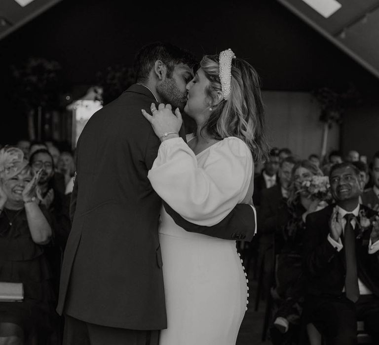 Bride wearing a chunky pearl headband kisses the groom for the first time as a married couple 