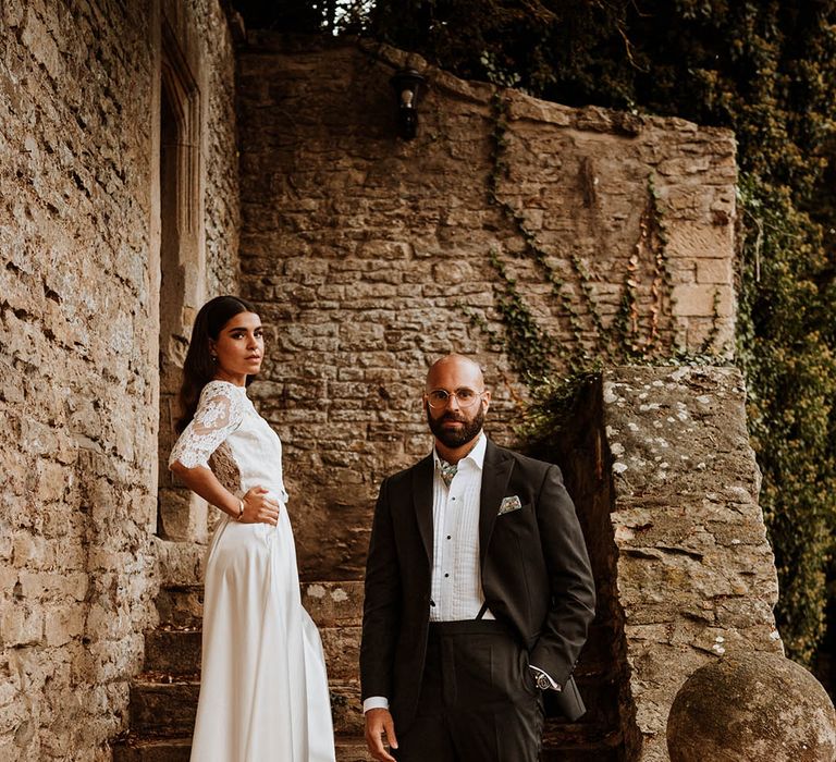 Bride in three quarter length sleeve wedding dress with lace detailing and full satin skirt posing with groom in classic black tuxedo with hand-painted silk cravat and handkerchief on the stairs of glasshouse wedding venue