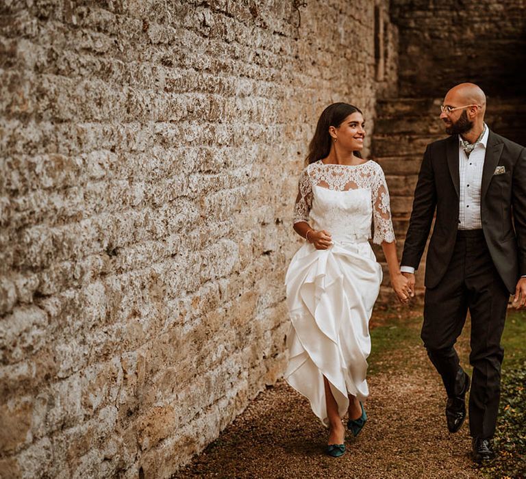 Groom in classic black tuxedo with hand-painted silk cravat and handkerchief and bride in three quarter length sleeve wedding dress with lace detailing and full satin skirt and bottle green satin wedding shoes holding hands and walking around wedding venue