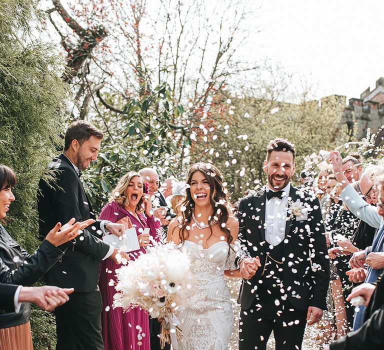 Bride in strapless boho fitted lace wedding dress with detachable sleeves having white confetti men with the groom in black tie 