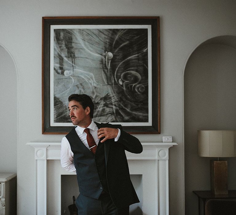 Groom wears three piece suit with maroon tie and puts on jacket on the morning of his wedding day