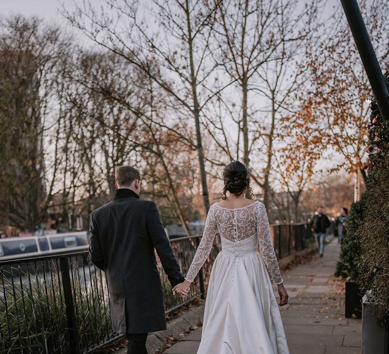 Bride in affeta material princess wedding dress with long lace sleeves  and cropped faux fur bridal cover up holding hands with groom in stylish black tuxedo with black bowtie walking around London