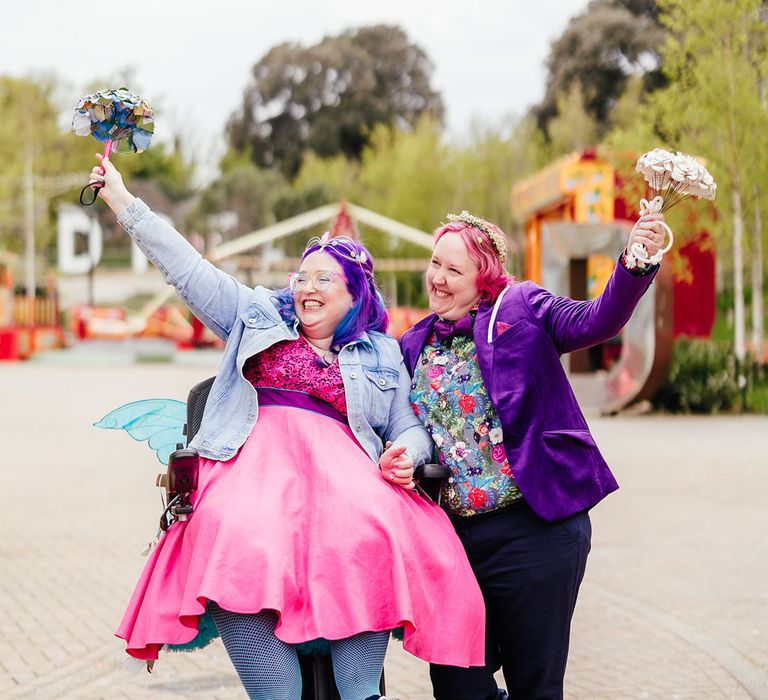 Bride in purple velvet blazer and black suit trousers with purple bowtie, pink patterned pocket square and colourful patterned shirt holding paper flower bouquet and bride in wheelchair wearing personalised denim jacket, pink wedding dress and blue tights holding colourful paper flower bouquet 