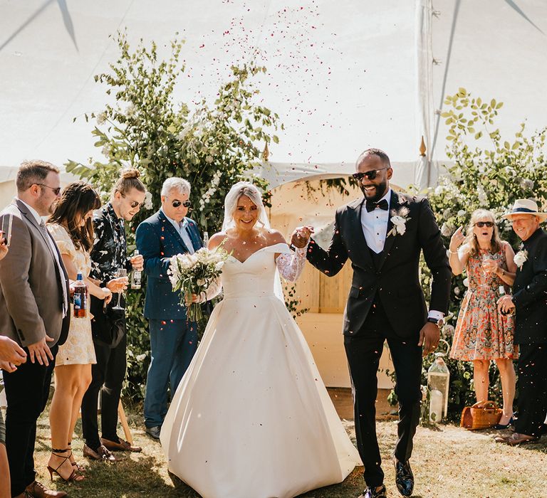 Bride in princess wedding dress and detachable lace sleeves walks through colourful confetti exit alongside her groom in black-tie