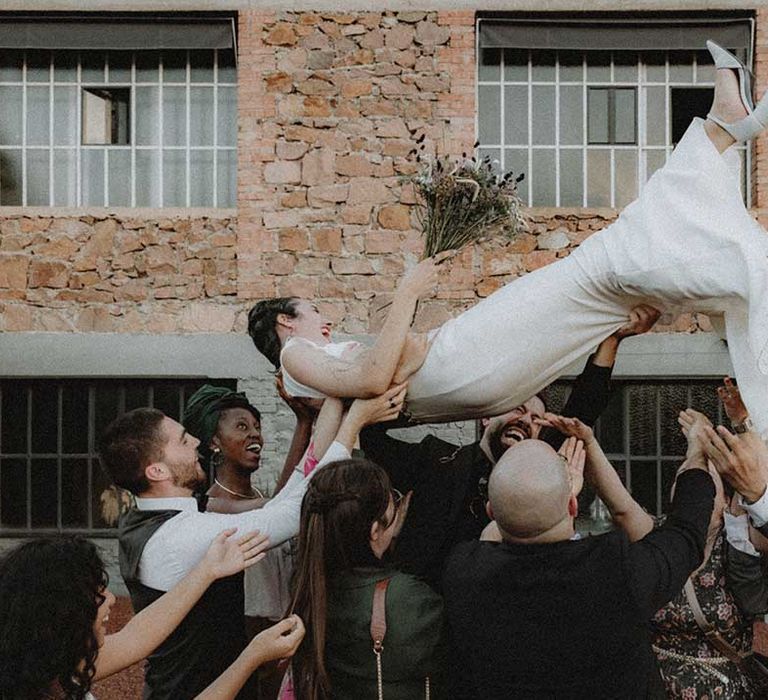 Lisa in pre-loved wedding dress is lifted by wedding guests outdoors 