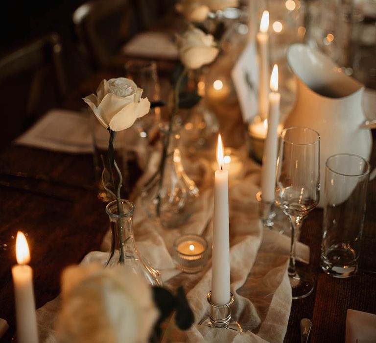 White wedding table runner with white candles and single white roses decorate the wedding table settings 